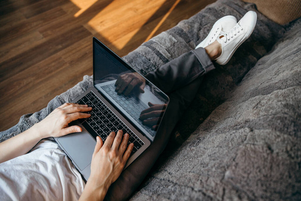 Красавчик пользуется ноутбуком, пока лежит на диване в гостиной с длинными ногами и белыми кроссовками
.  - Фото, изображение