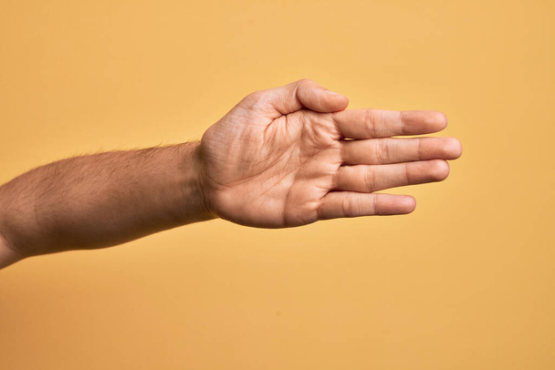 Ręka białego młodzieńca pokazuje palce nad odizolowanym żółtym tłem rozciągającym się i sięgającym z otwartą dłonią po uścisk dłoni, pokazując dłoń - Zdjęcie, obraz