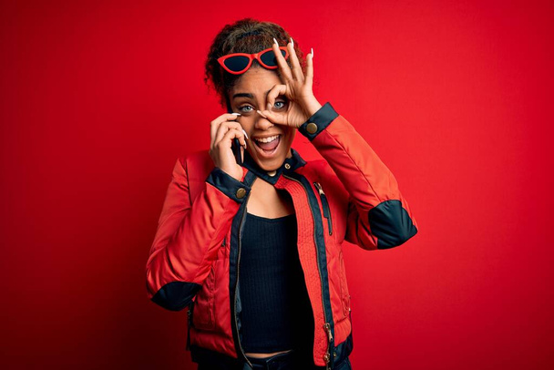 Genç Afrikalı Amerikalı kız kırmızı arka planda akıllı telefondan konuşuyor. Gülümsüyor. El ele verip parmaklarının arasından bakıyor. - Fotoğraf, Görsel