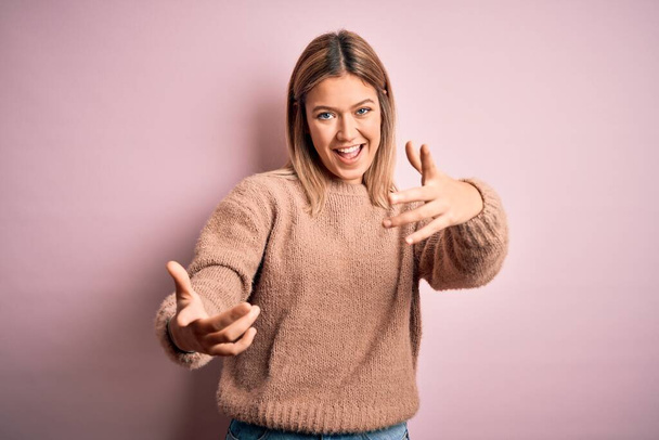 Young beautiful blonde woman wearing winter wool sweater over pink isolated background smiling cheerful offering hands giving assistance and acceptance. - Фото, изображение