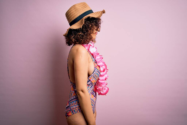 Mujer árabe hermosa joven de vacaciones con traje de baño y flores de lei hawaiano mirando a un lado, pose de perfil relajante con cara natural con sonrisa confiada
. - Foto, Imagen