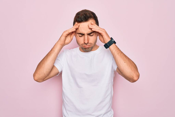 Beau homme aux yeux bleus portant un t-shirt blanc décontracté debout sur fond rose avec la main sur la tête pour la douleur à la tête parce que le stress. Migraine souffrante
. - Photo, image
