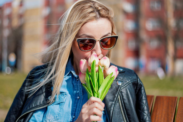 Hermosa mujer rubia en un paño casual oliendo tulipanes al aire libre, disfrutando del olor de las flores en el día de primavera
. - Foto, Imagen