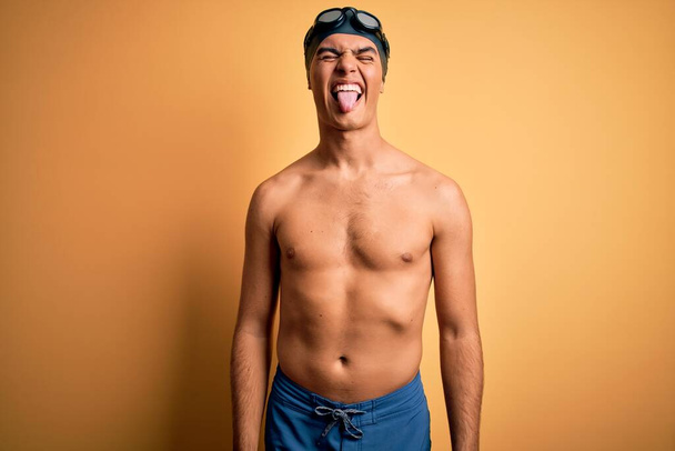 Νεαρός όμορφος άντρας χωρίς πουκάμισο φορώντας μαγιό και σκουφάκι κολύμβησης πάνω από απομονωμένο κίτρινο φόντο που βγάζει τη γλώσσα του χαρούμενη με την αστεία έκφραση. Έννοια συγκίνησης. - Φωτογραφία, εικόνα