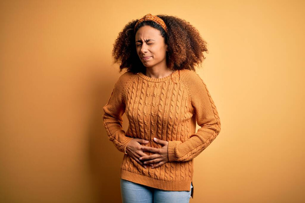 Mujer afroamericana joven con cabello afro que usa suéter casual sobre fondo amarillo con la mano en el estómago porque la indigestión, la enfermedad dolorosa sensación de malestar. Concepto Ache
. - Foto, imagen