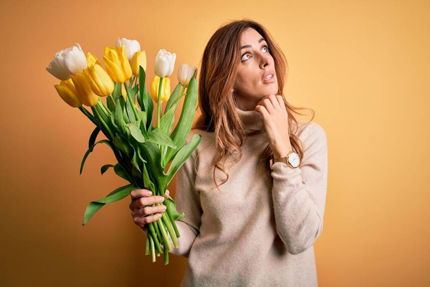 Young beautiful brunette woman holding bouquet of yellow tulips over isolated background with hand on chin thinking about question, pensive expression. Smiling with thoughtful face. Doubt concept. - Photo, Image