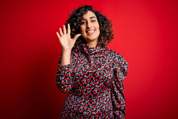 Молодая красивая кудрявая арабская женщина в обычном цветочном платье, стоящем на красном фоне, показывая пальцами номер пять, улыбаясь уверенно и счастливо
. - Фото, изображение