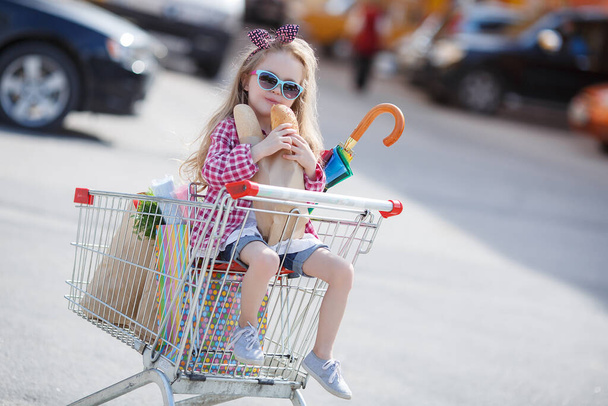 Adorable baby kid with trolley choosing fresh vegetables in local store. Girl with a big grocery cart in a parking lot near a store. A little girl with a big trolley buys fresh food and vegetables in a big supermarket.Little housewife with groceries - Foto, Imagem