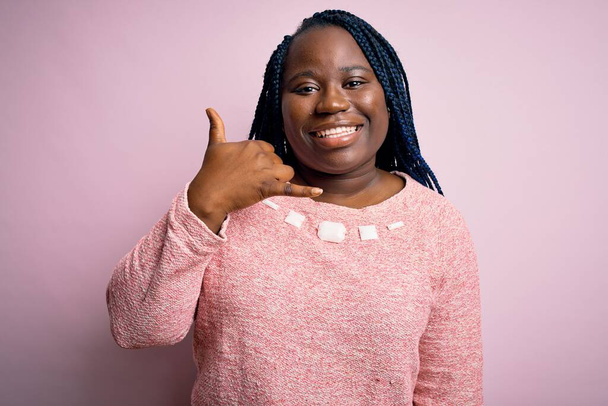 Afroamerykanka plus rozmiar kobieta z warkoczami w swobodnym swetrze na różowym tle uśmiechając się wykonując telefon gestem z ręki i palców jak rozmawiając przez telefon. Pojęcia komunikacyjne. - Zdjęcie, obraz