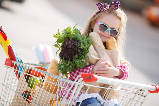 Roztomilé dítě s vozíkem vybírá čerstvou zeleninu v místním obchodě. Dívka s velkým nákupním vozíkem na parkovišti poblíž obchodu. Malá holka s velkým vozíkem kupuje čerstvé jídlo a zeleninu ve velkém supermarketu.Malá hospodyňka s potravinami - Fotografie, Obrázek