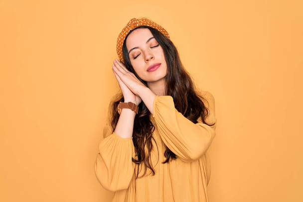 Young beautiful woman wearing casual t-shirt and diadem standing over yellow background sleeping tired dreaming and posing with hands together while smiling with closed eyes. - Photo, image