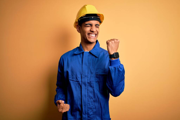 Joven trabajador afroamericano guapo vestido con uniforme azul y casco de seguridad celebrando sorprendido y sorprendido por el éxito con los brazos levantados y los ojos cerrados. Concepto ganador
. - Foto, Imagen