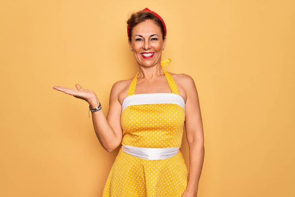 中年シニアピンアップ女性身に着けています50スタイルレトロドレス上の黄色の背景笑顔陽気なプレゼンテーションとポインティングで手見カメラ. - 写真・画像