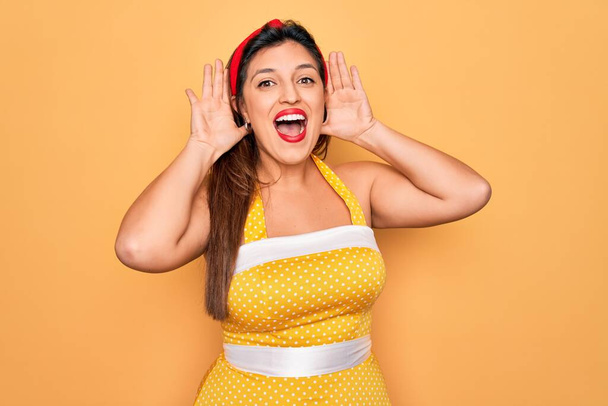 Молода іспаномовна жінка в модному стилі 50-х років над жовтим тлом Усміхнений веселий граючи зазирнути в бу з руками, що показують обличчя. Здивований і вийшов
 - Фото, зображення