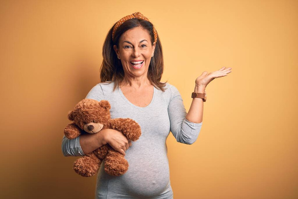 Mulher grávida de meia-idade esperando bebê segurando ursinho pelúcia animal muito feliz e animado, expressão vencedora celebrando vitória gritando com grande sorriso e mãos levantadas
 - Foto, Imagem