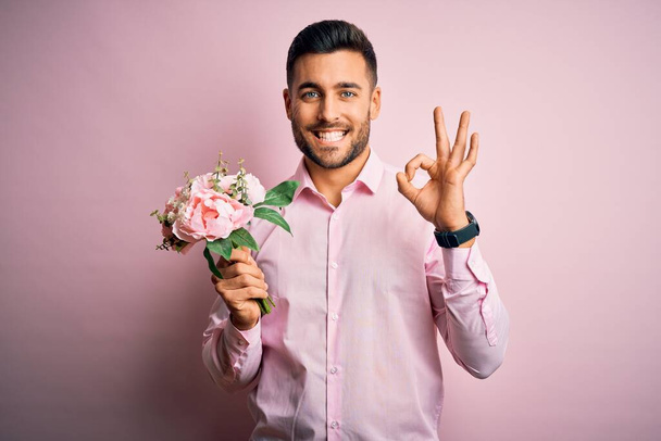 Νέοι romatic άνθρωπος κρατώντας μπουκέτο από λουλούδια άνοιξη πάνω από το ροζ απομονωμένο φόντο κάνει ok υπογράψει με τα δάχτυλα, εξαιρετικό σύμβολο - Φωτογραφία, εικόνα