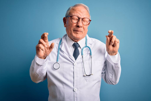 Старший седой врач в стетоскопе и медицинском пальто на синем фоне жестом скрещенном пальце улыбается с закрытыми глазами и надеждой. Удача и суеверие
. - Фото, изображение