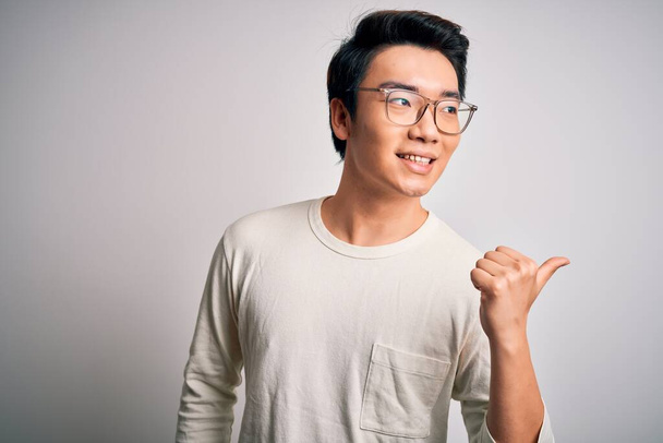 Genç, yakışıklı Çinli bir adam beyaz arka planda günlük tişört ve gözlük takıyor. Gülümsüyor, gülümsüyor ve başparmağıyla yana bakıyor.. - Fotoğraf, Görsel