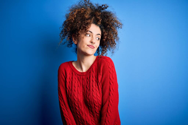 Jonge mooie vrouw met krullend haar en piercing dragen casual rode trui glimlachen kijken naar de zijkant en staren weg denken. - Foto, afbeelding