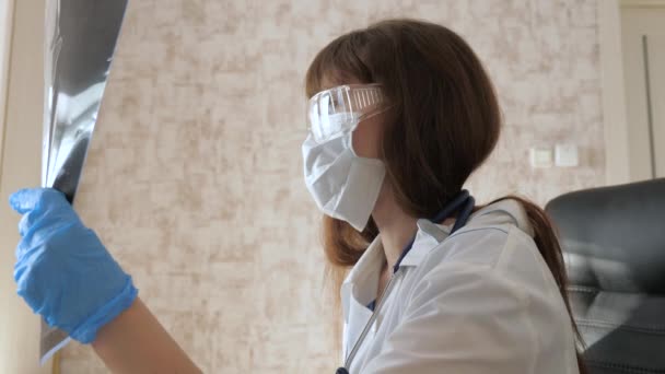 Μια γυναίκα γιατρός σε ένα γραφείο εξετάζει μια ακτινογραφία του ασθενούς στο νοσοκομείο. Ο γιατρός δουλεύει στο νοσοκομείο με λάπτοπ. Ιατρική περίθαλψη και υγειονομική περίθαλψη έννοια. πανδημία του κορονοϊού, πνευμονία COVID-19 - Πλάνα, βίντεο