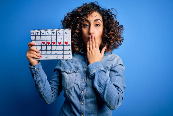Junge schöne, lockige arabische Frau, die einen Zeitrahmen-Kalender hält, der den Menstruationszyklus kontrolliert, Mund mit der Hand vor Scham über Fehler geschockt, Ausdruck von Angst, Angst in der Stille, geheimes Konzept - Foto, Bild