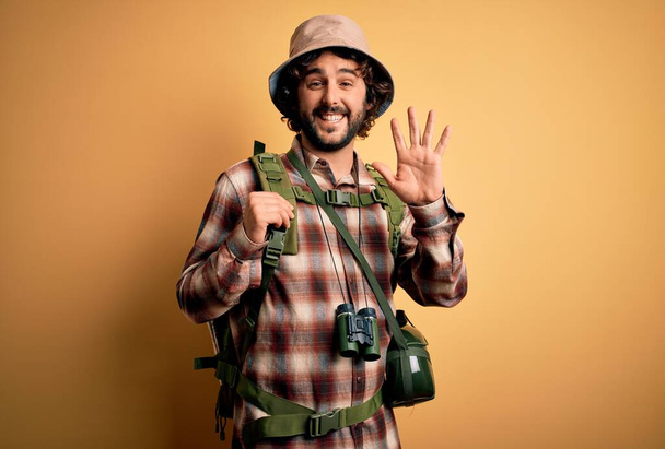 Giovane escursionista uomo con capelli ricci e barba escursionismo indossa zaino e borraccia d'acqua rinuncia dicendo ciao felice e sorridente, amichevole gesto di benvenuto
 - Foto, immagini
