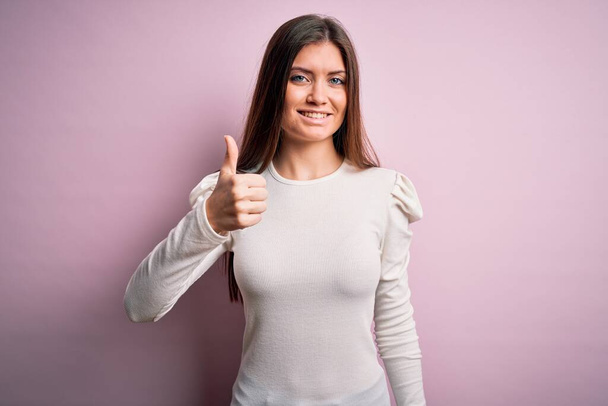 Νεαρή όμορφη γυναίκα με μπλε μάτια φορώντας casual λευκό t-shirt πάνω από ροζ φόντο κάνει ευτυχισμένη αντίχειρες επάνω χειρονομία με το χέρι. Έγκριση έκφρασης κοιτάζοντας την κάμερα που δείχνει επιτυχία. - Φωτογραφία, εικόνα