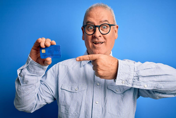 Człowiek w średnim wieku hoary nosi okulary trzymając kartę kredytową jako pieniądze do zapłaty bardzo szczęśliwy wskazując ręką i palcem - Zdjęcie, obraz