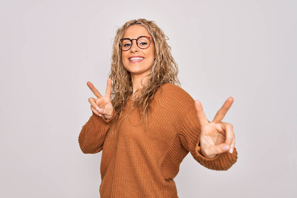 Young beautiful blonde woman wearing casual sweater and glasses over white background smiling looking to the camera showing fingers doing victory sign. Number two. - Photo, Image
