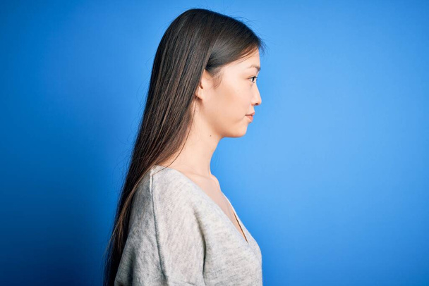 Joven hermosa mujer asiática con suéter casual de pie sobre fondo aislado azul mirando a un lado, pose de perfil de relax con cara natural y sonrisa confiada
. - Foto, imagen