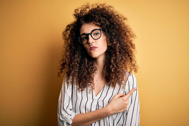 Young beautiful woman with curly hair and piercing wearing striped shirt and glasses Pointing with hand finger to the side showing advertisement, serious and calm face - Photo, Image