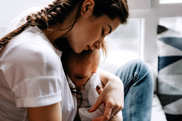 young pretty woman breastfeeding her little daughter sitting on the window sill of her bedroom - Foto, afbeelding
