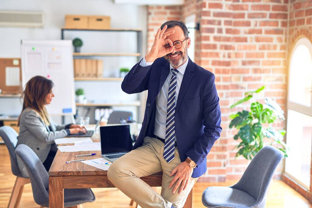 Przystojny biznesmen w średnim wieku, noszący okulary siedzący na biurku w biurze, wykonujący gest ok z uśmiechem dłoni, okiem patrzącym przez palce ze szczęśliwą twarzą. - Zdjęcie, obraz