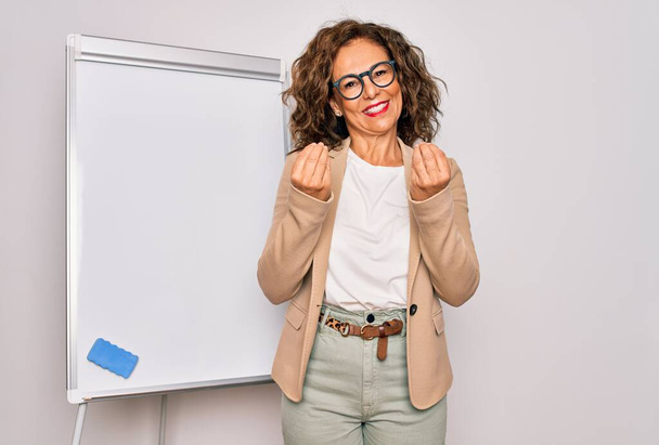 Middle age senior business woman standing on seminar presentation by magnectic blackboard doing money gesture with hands, asking for salary payment, millionaire business - Photo, Image