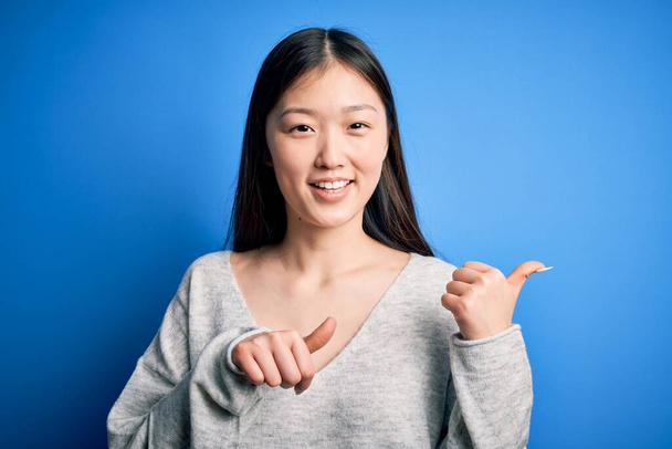 Jovem bela mulher asiática vestindo camisola casual em pé sobre azul isolado fundo Apontando para trás com a mão e polegares para cima, sorrindo confiante
 - Foto, Imagem