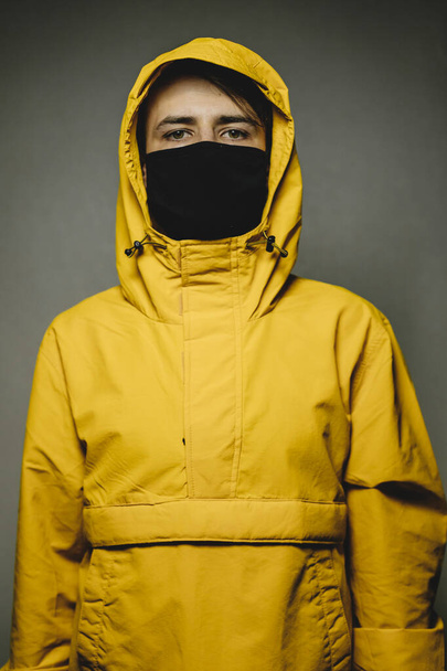 young skinny man wearing a yellow hooded jacket and black face mask with an aggressive serious look - Zdjęcie, obraz