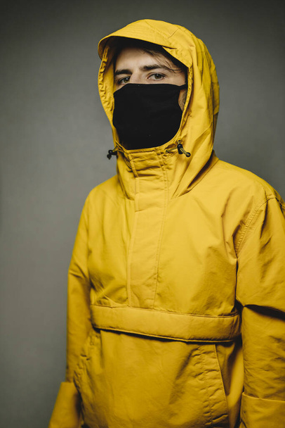young skinny man wearing a yellow hooded jacket and black face mask with an aggressive serious look - Foto, Bild