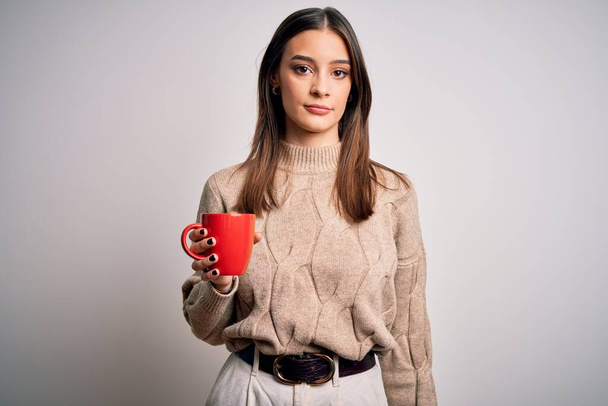 Giovane bella donna bruna bere tazza rossa di caffè su sfondo bianco isolato con un'espressione fiduciosa sul viso intelligente pensando serio
 - Foto, immagini