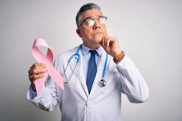 Średniego wieku przystojny siwowłosy lekarz trzymający różową wstążkę raka poważna twarz myśląc o pytaniu, bardzo mylący pomysł - Zdjęcie, obraz