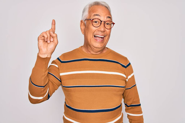 Middle age senior grey-haired handsome man wearing glasses and casual striped sweater pointing finger up with successful idea. Exited and happy. Number one. - Photo, Image