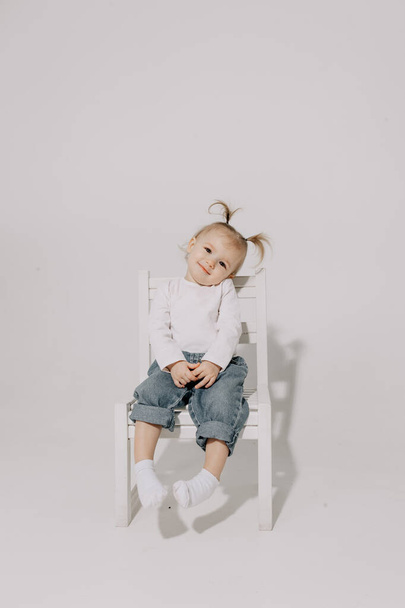 cute little girl with a ponytail hairstyle dressed in a white T-shirt and denim pants posing for a photo on a white background in a photo studio - Fotografie, Obrázek