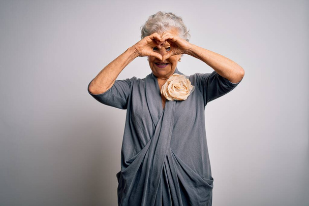 Senior beautiful grey-haired woman wearing casual dress standing over white background Doing heart shape with hand and fingers smiling looking through sign - Foto, Bild