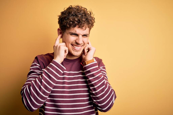 Ein junger blonder, gutaussehender Mann mit lockigem Haar trägt einen lässig gestreiften Pullover, der die Ohren mit Fingern bedeckt, die sich über den Lärm lauter Musik ärgern. Gehörloses Konzept. - Foto, Bild