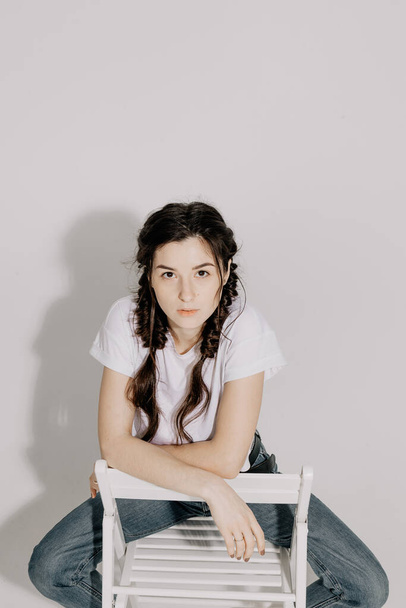 Young pretty woman with pigtail braid dressed in white t-shirt and jeans posing for photo on a white background in front of studio light - 写真・画像