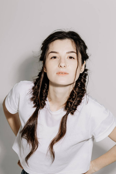 Young pretty woman with pigtail braid dressed in white t-shirt and jeans posing for photo on a white background in front of studio light - Фото, зображення