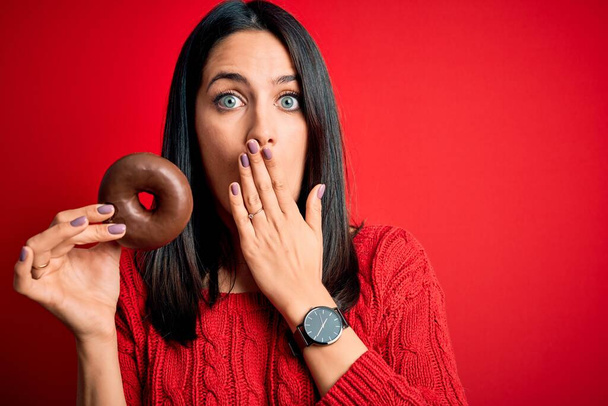 Молода брюнетка з блакитними очима їсть шоколадний пончик над червоним ізольованим фоновим покривом ротом з рукою, шокованим соромом за помилку, вираження страху, наляканий тишею, таємна концепція
 - Фото, зображення