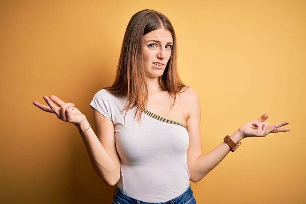Young beautiful redhead woman wearing casual t-shirt over isolated yellow background clueless and confused with open arms, no idea concept. - Photo, Image