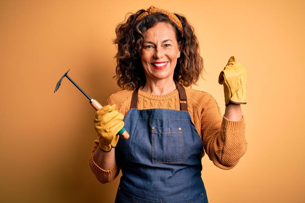 Femme jardinière du Moyen Âge portant tablier tenant râteau sur fond jaune isolé criant fier et célébrant la victoire et le succès très excité, acclamant l'émotion
 - Photo, image