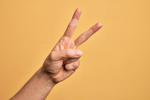 Hand eines kaukasischen jungen Mannes mit Fingern über einem isolierten gelben Hintergrund, der die Zahl 2 zählt, die zwei Finger zeigt, Sieg und Siegersymbol gestikuliert - Foto, Bild