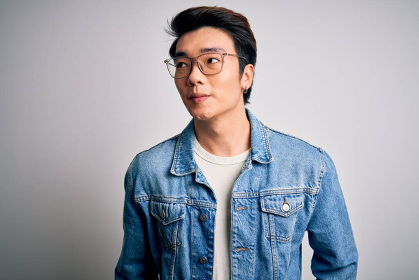 Young handsome chinese man wearing denim jacket and glasses over white background smiling looking to the side and staring away thinking. - Foto, Imagem